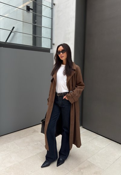 Mujer con abrigo largo marrón, camisa blanca, vaqueros oscuros, botas negras puntiagudas y gafas de sol, de pie contra una pared gris en el interior.