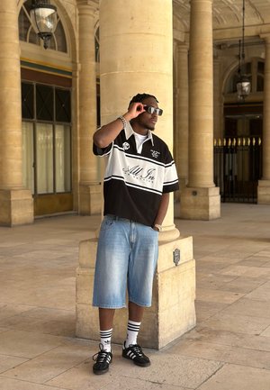 Hombre con gafas de sol, lleva un polo blanco y negro y pantalones cortos vaqueros, apoyado en una columna de piedra en el pasillo arqueado de un edificio histórico.