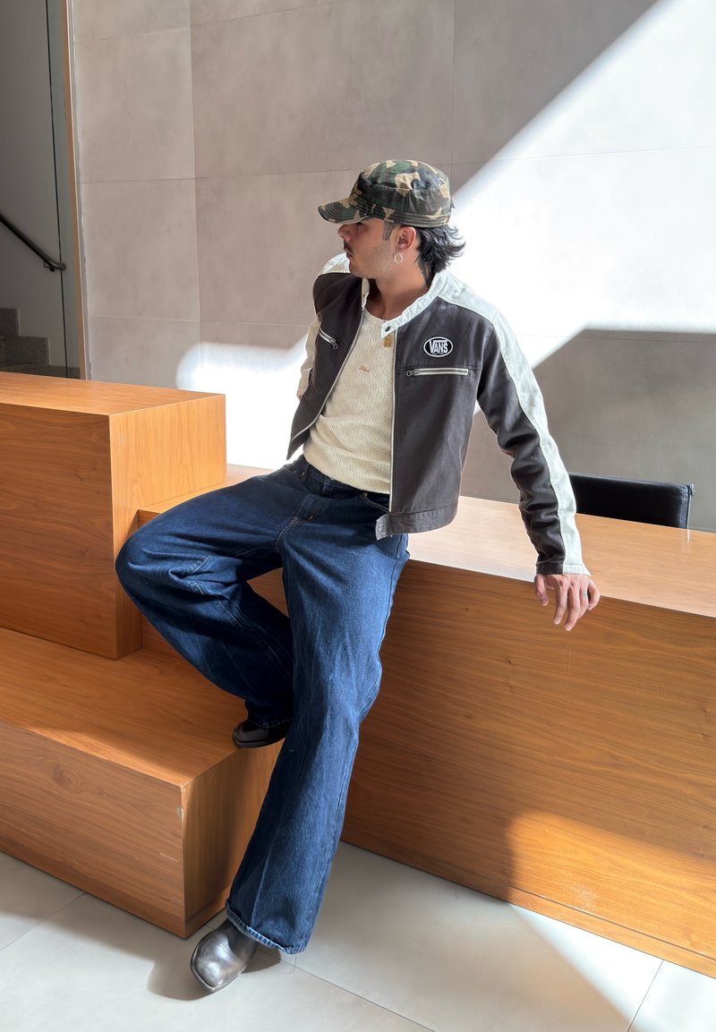 Uomo con cappello camouflage, giacca Vans, maglione bianco e jeans seduto su gradini di legno in un moderno interno illuminato dal sole.