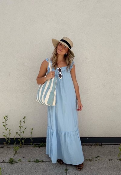Vestido maxi a rayas azul claro con tirantes de hombro con volantes y falda de capas. Accesorios incluyen un bolso de mano a rayas y un sombrero de ala ancha.