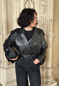 Veste en cuir noir coupée, à double boutonnage, avec épaulettes, taille ceinturée et manches larges, associée à un jean noir taille haute et un sac noir.