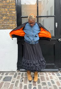 Denim bomberjakke med orange fôr over et svart lagvis volangskjørt og brune støvler. Brosteinsbelagt bakke, svart dør i bakgrunnen.