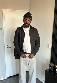 Chemise noire à boutons portée sur un t-shirt blanc, pantalon de jogging gris et bonnet noir. Debout avec un sac, mains à la taille dans un cadre intérieur simple.