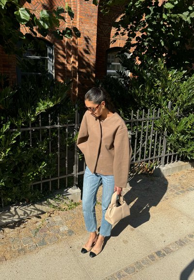 Mujer con chaqueta beige y vaqueros azules, sosteniendo un bolso beige, de pie en la acera cerca de una valla con plantas verdes y un edificio de ladrillos bajo la luz del sol.