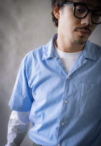 Cămașă cu dungi albastre și albe, cu mâneci scurte, guler, închidere cu nasturi în față și un singur buzunar pe piept. Este purtată peste un maiou alb.