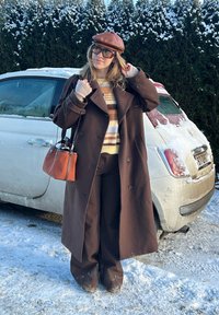 Femme portant un long manteau marron, un pull rayé et un chapeau, debout dans la neige près d'une voiture blanche recouverte de neige.