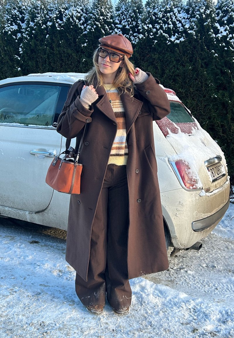 Femme portant un long manteau marron, un pull rayé et un chapeau, debout dans la neige près d'une voiture blanche recouverte de neige.