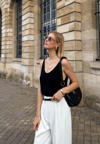 Schwarzes ärmelloses Oberteil, weiße weit geschnittene Hosen, schwarze Schultertasche, goldene Ohrringe und Armbänder; stehend vor einer Steinmauer.