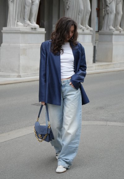 Mujer con cabello rizado que lleva una chaqueta azul, top blanco, vaqueros anchos claros y zapatos blancos, sosteniendo un bolso azul, caminando por la acera cerca de estatuas.