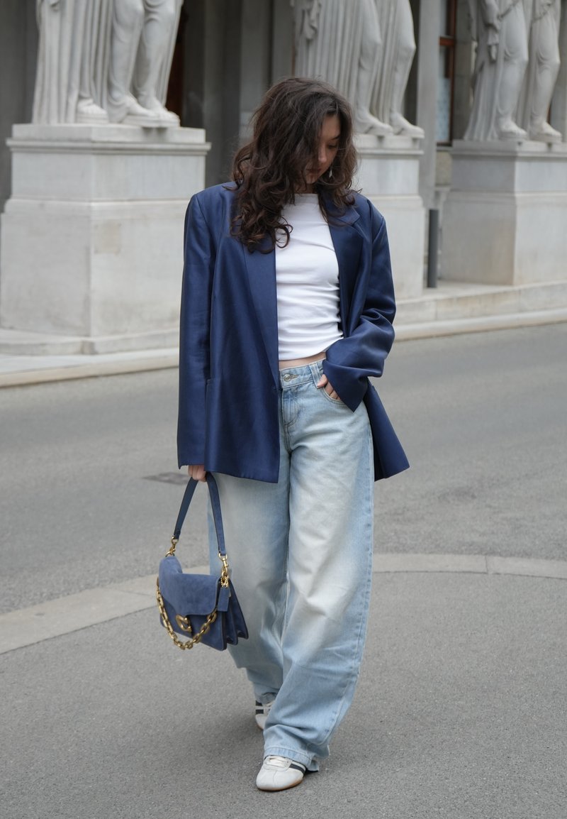 Γυναίκα με σγουρά μαλλιά, που φορά μπλε σακάκι, λευκό τοπ, φαρδιά ανοιχτόχρωμα τζιν και λευκά παπούτσια, κρατώντας μπλε τσάντα, περπατά στο πεζοδρόμιο κοντά σε αγάλματα.