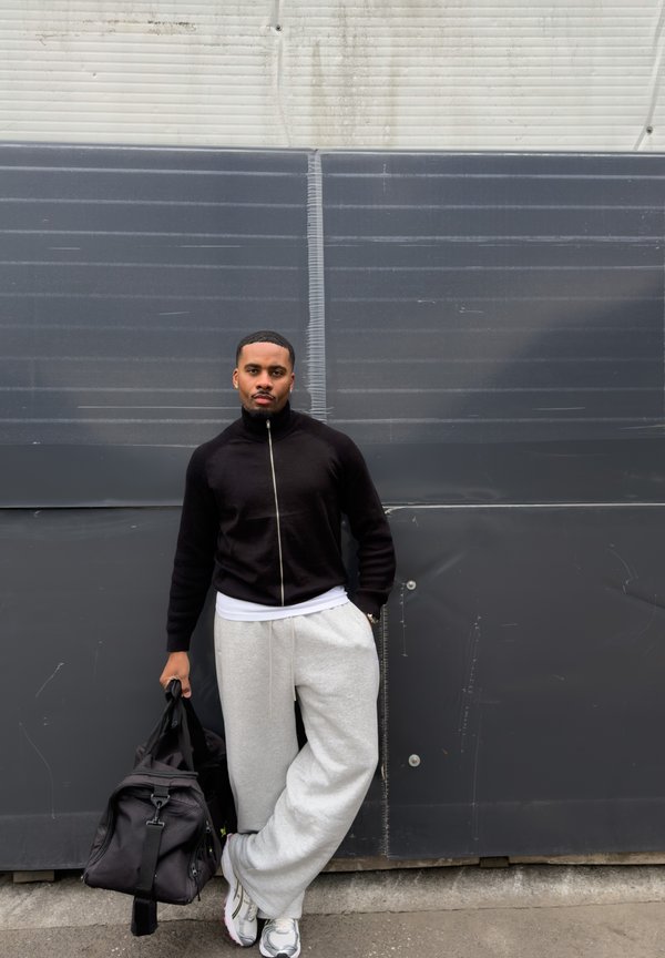 Homme en veste noire zippée et pantalon de jogging gris appuyé contre un mur sombre, tenant un sac de sport noir d'une main.