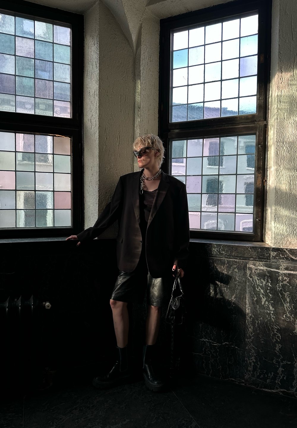 Μαύρη oversized σαλοπέτα πάνω από σκούρο τοπ, συνδυασμένη με γυαλιστερά μαύρα σορτς. Μαύρες μπότες με χοντρή �σόλα, κρατώντας μια υφασμάτινη τσάντα, στέκεται δίπλα σε παράθυρα.