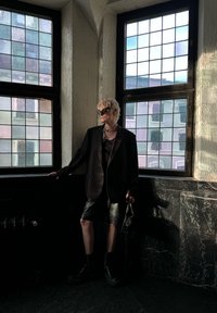 Μαύρη oversized σαλοπέτα πάνω από σκούρο τοπ, συνδυασμένη με γυαλιστερά μαύρα σορτς. Μαύρες μπότες με χοντρή σόλα, κρατώντας μια υφασμάτινη τσάντα, στέκεται δίπλα σε παράθυρα.