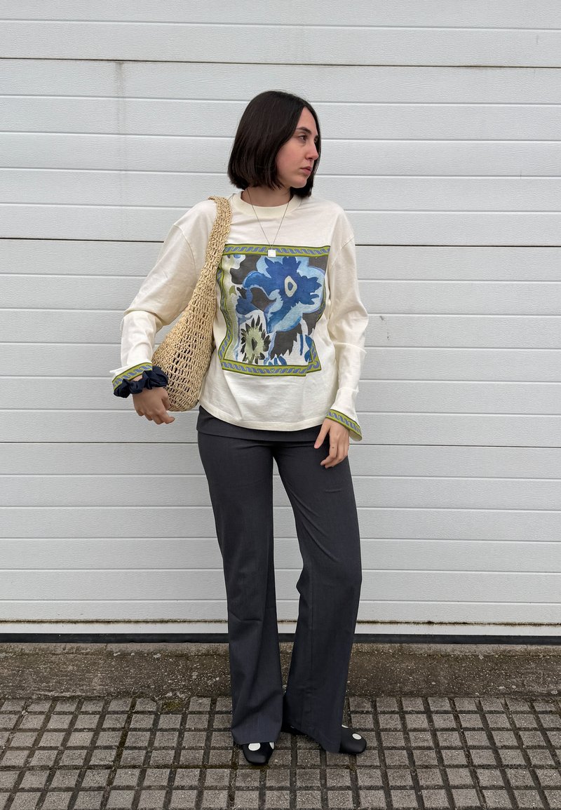 Femme aux cheveux courts et foncés portant un pull crème avec un imprimé fleuri bleu, un pantalon noir, des chaussures noires et portant un sac à épaule tissé.