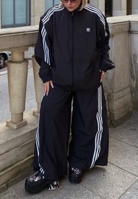 Μαύρο αθλητικό μπουφάν και φαρδιά παντελόνια με τρεις λευκές ρίγες, συνδυασμένα με πλατφόρμες ξυλοτύπου που διαθέτουν σχέδιο φλόγας. Εξωτερικός χώρος.