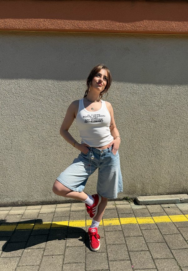 Jonge vrouw draagt een wit tanktop met tekst koffie, spijkershorts en rode sneakers, staand op een stoep tegen een beige muur.