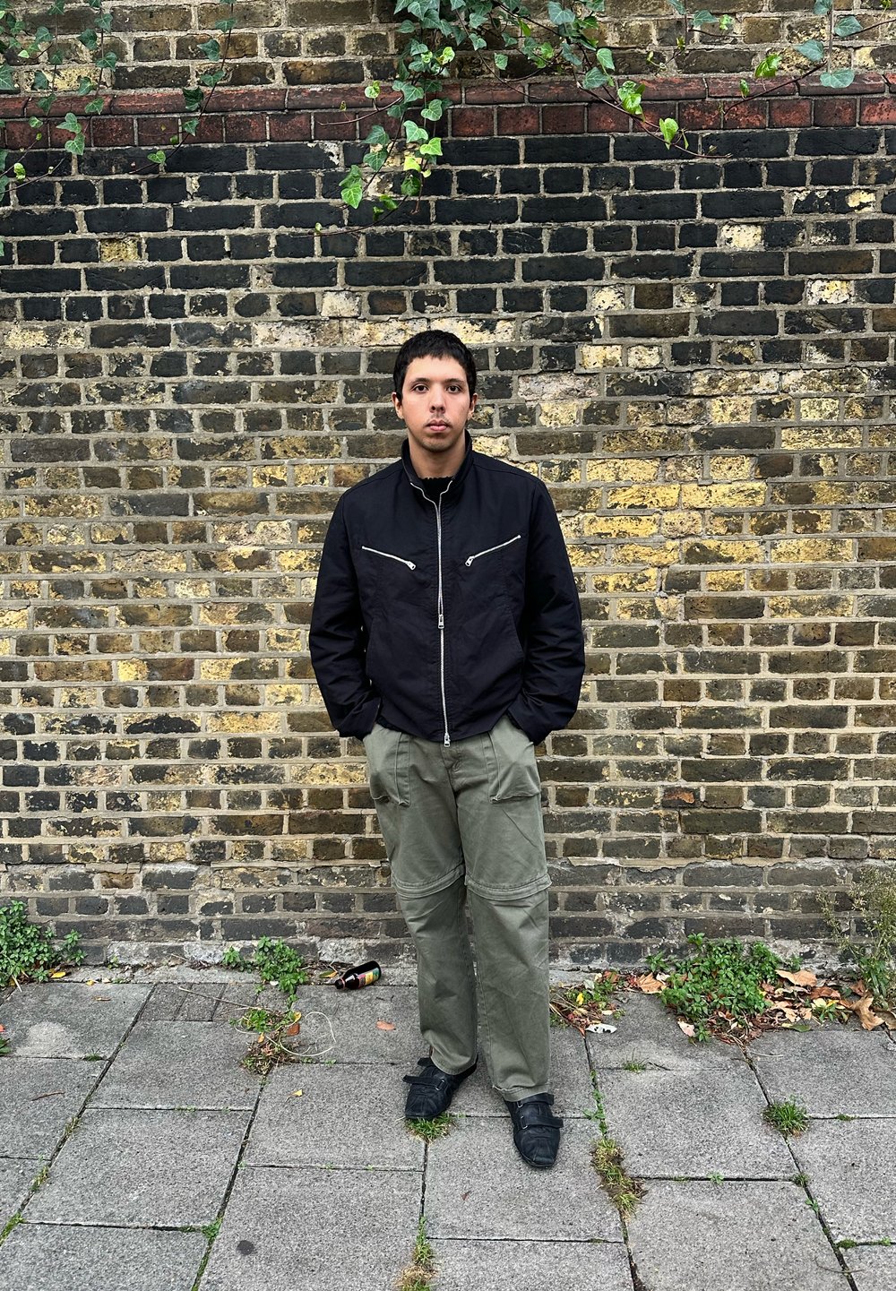 Crna jakna na zip s srebrnim detaljima, maslinasto zelene cargo hlače i crne cipele, stoji naslonjena na strukturiranu ciglanu zid.