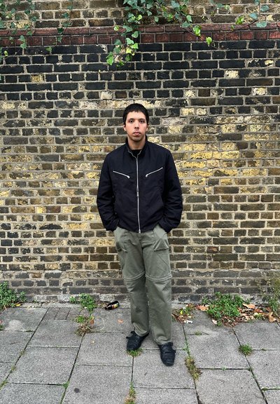 Chaqueta negra con cremallera y herrajes plateados, pantalones cargo verde oliva y zapatos negros, de pie frente a una pared de ladrillos texturizada.