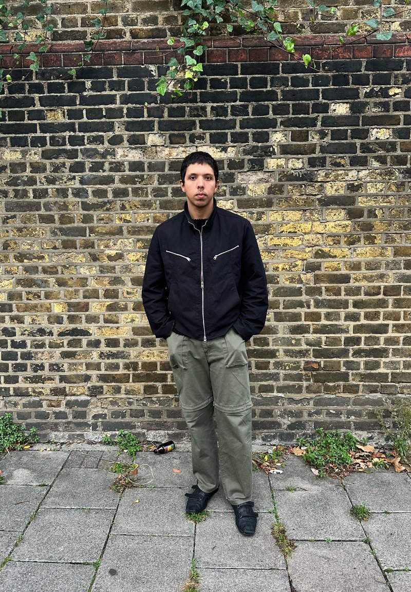 Fekete cipzáras dzseki ezüst hardware-rel, olíva zöld cargo nadrág és fekete cipő, egy textúrált tégla fal előtt áll.