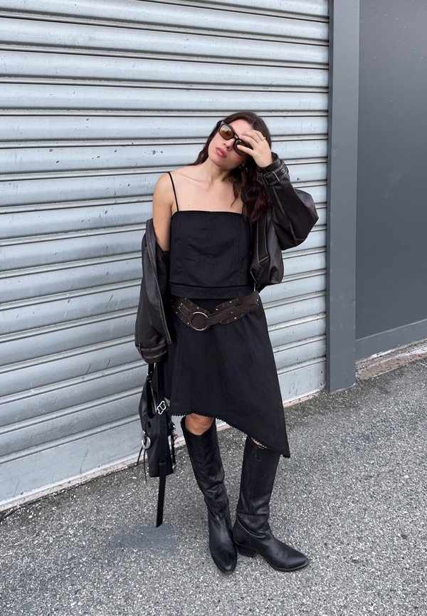 Femme debout près d'un volet métallique portant des lunettes de soleil, une robe noire sans manches, une ceinture marron, une veste en cuir noire drapée sur les épaules et des bottes montantes.
