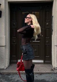 Black sheer long-sleeve top, leather mini skirt, thigh-high black boots, and a red handbag. Standing in front of a dark wooden door.