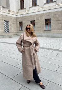 Beige, textured wool coat with wide lapels and a tied waist, paired with dark jeans and brown shoes. Outdoor setting with smooth pavement.