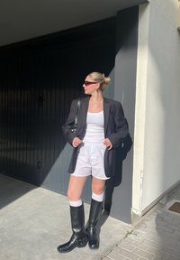 Μαύρο σακάκι πάνω από λευκή μπλούζα και λευκά σορτς, συνδυασμένο με μαύρες μπότες μέχρι το γόνατο και λευκές κάλτσες. Συμπεριλαμβάνονται γυαλιά ηλίου και τσάντα ώμου.