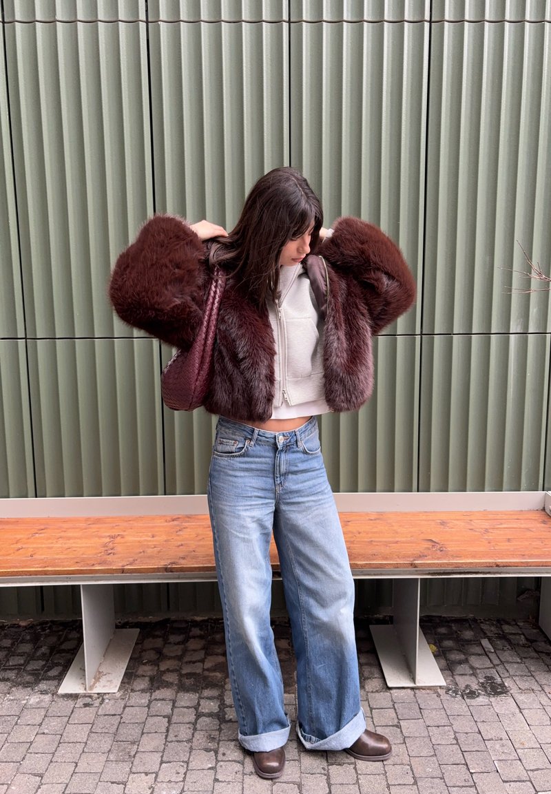 Femme ajustant une veste en fourrure bordeaux, portant un hoodie blanc court, un jean à jambes larges, des bottes marron et tenant un sac assorti bordeaux près d'un mur vert et d'un banc en bois.