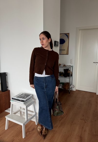 Cardigan marrón de pelaje sobre una blusa blanca, combinado con una falda de mezclilla larga y botas puntiagudas marrones. Sosteniendo un bolso verde. Suelo de madera clara.