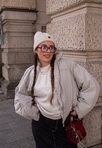 Casaco puffer cinza claro, sweatshirt branca, saia preta riscada, gorro de malha creme com um coração vermelho, óculos roxos, bolsa vermelha com detalhes.