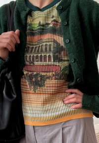 Cardigan felpudo verde escuro com fechos de botão sobre um top canelado multicolorido com uma impressão de paisagem. Calças de tecido cinzentas e uma bolsa preta visíveis.