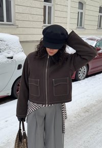 Veste marron courte avec coutures blanches et deux poches avant, portée par-dessus une chemise rayée. Pantalon gris à jambes larges et chapeau noir. Fond enneigé.