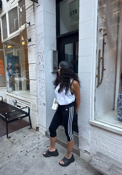 Leggings negros de adidas con rayas blancas, una camiseta sin mangas blanca, una gorra negra y sandalias de plataforma negras. De pie contra una pared cerca de una tienda.