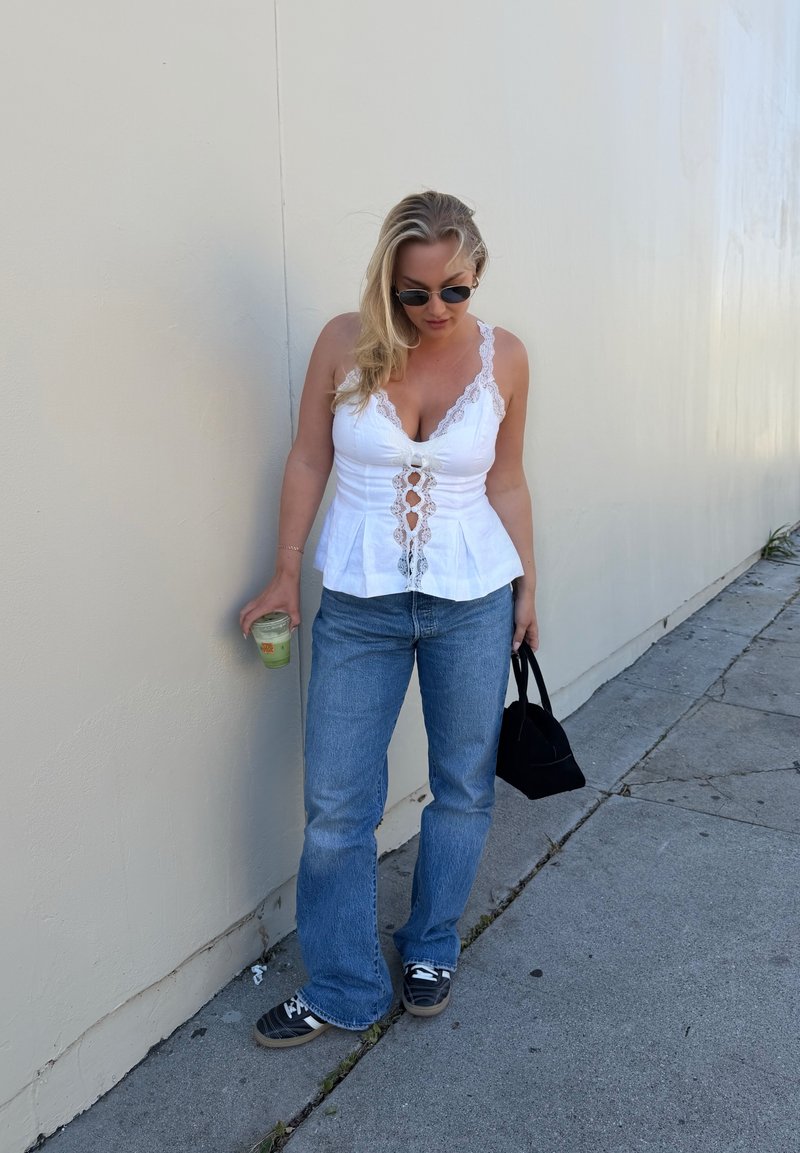 Donna con top di pizzo bianco e jeans blu appoggiata a un muro, tiene in una mano una bevanda e nell'altra una borsa nera.