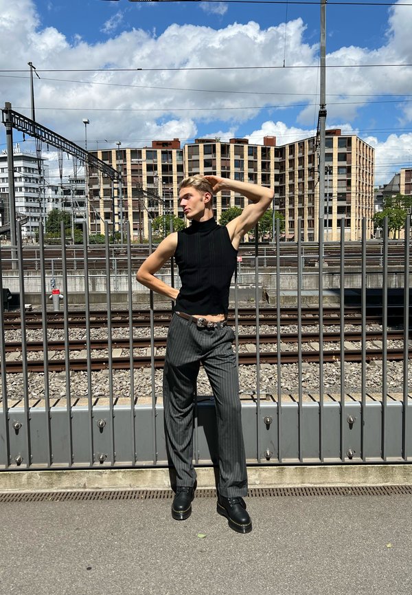 Juodas rankovių neturintis golfas su vertikaliomis briaunomis; pilki dryžuoti kelnės; masyvūs juodi batai; miesto fonas su metaline tvora ir pastatais.