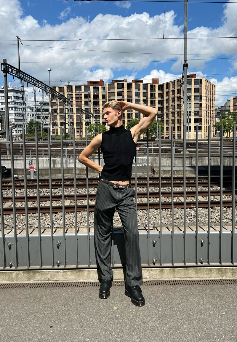 Crni top bez rukava s visokim ovratnikom i okomitim rebrastim uzorkom; sive hlače s uskim prugama; masivne crne cipele; urbani ambijent s metalnom ogradom i zgradama.