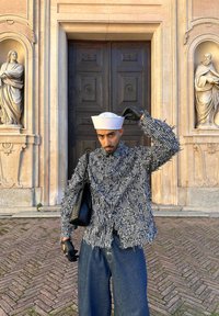 Homme portant une veste à motifs, un chapeau de marin blanc et des gants, ajustant son chapeau, debout devant une grande porte en bois ornée encadrée par des statues en pierre.