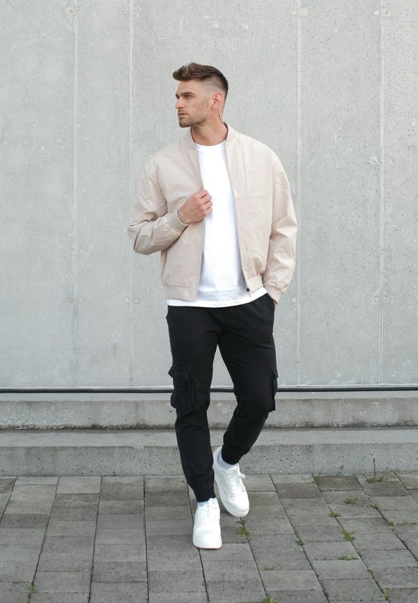 Mand gående på belagt område iført beige jakke, hvid skjorte, sorte cargobukser og hvide sneakers med en betonvæg i baggrunden.