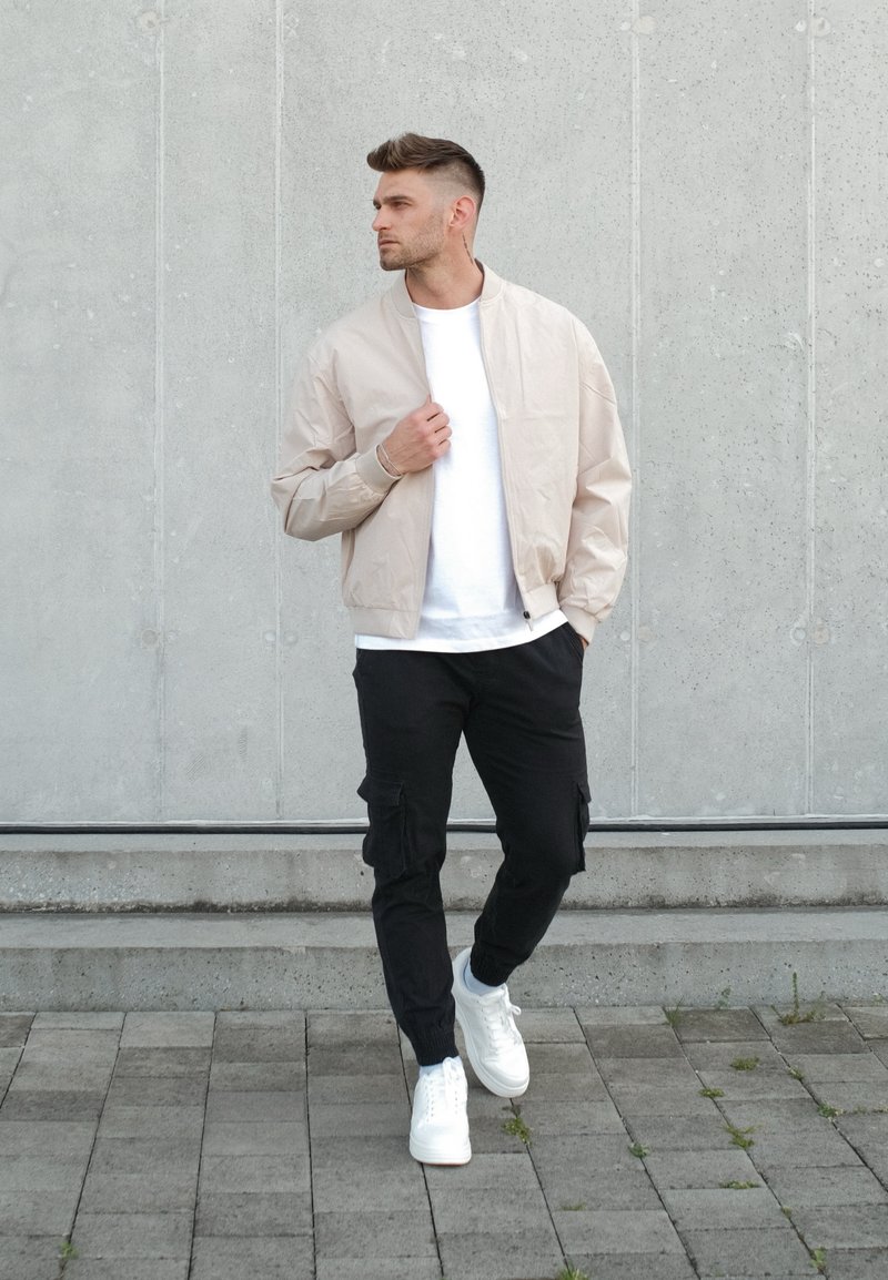 Homme marchant sur une zone pavée portant une veste beige, une chemise blanche, un pantalon cargo noir et des baskets blanches devant un mur en béton.