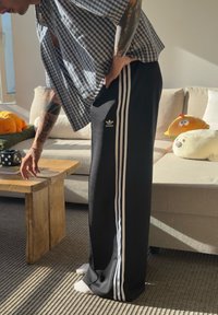 Calças de desporto pretas com três riscas brancas nas laterais, feitas de um tecido macio. Usadas com uma camisa larga de padrão xadrez, com as mãos repousadas numa mesa de madeira.