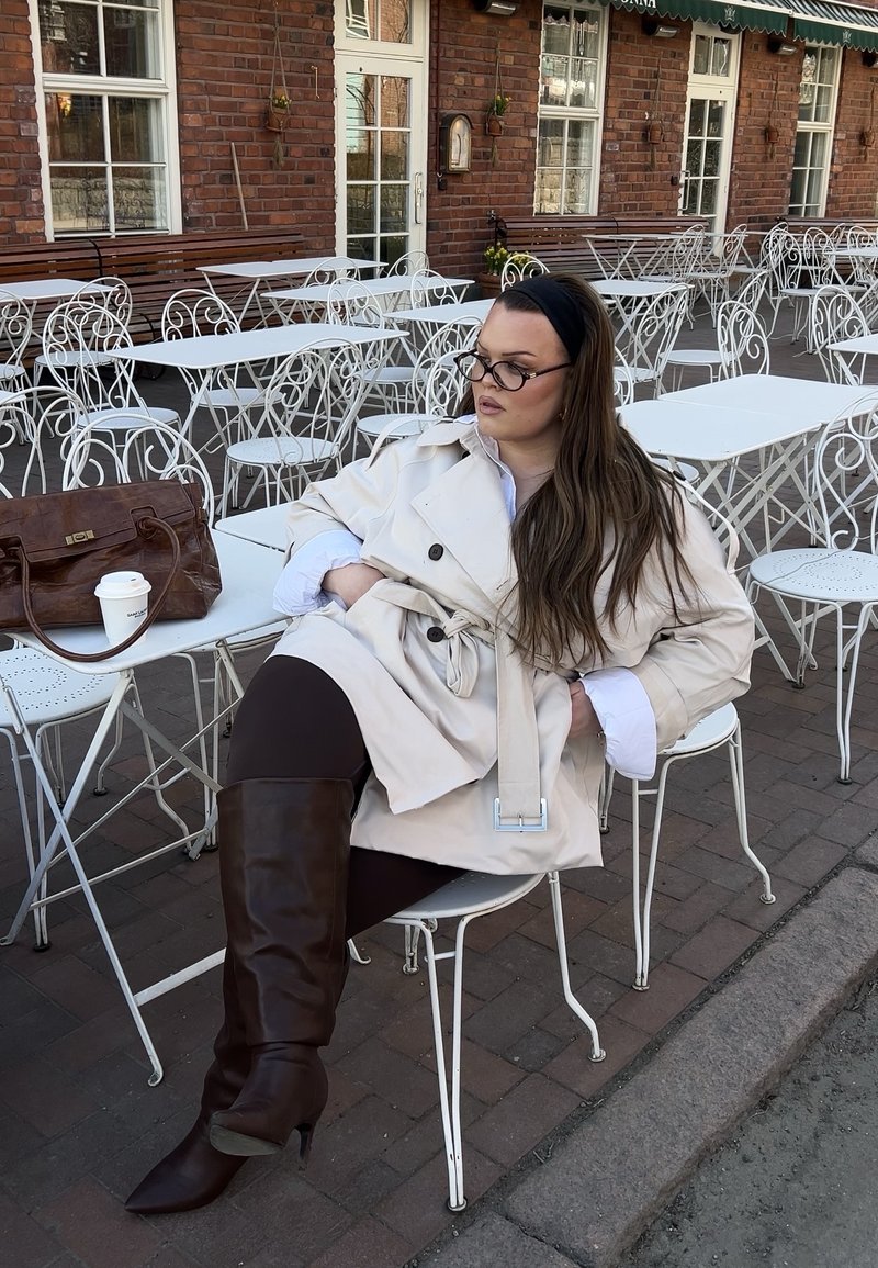 Femme portant un manteau beige et des bottes marron assise dans un café en plein air avec des tables et des chaises en métal blanc sur un sol pavé de briques.