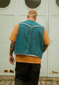 Veste en jean avec une teinte bleue, bords effilochés, portée sur une chemise orange côtelée. Jean noir avec des détails de surpiqûres discrètes. Vue de dos.