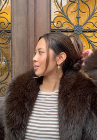 Abrigo de piel marrón con textura gruesa, camisa a rayas blancas y grises, y pendientes en forma de corazón. El fondo muestra un diseño ornamentado de hierro forjado.