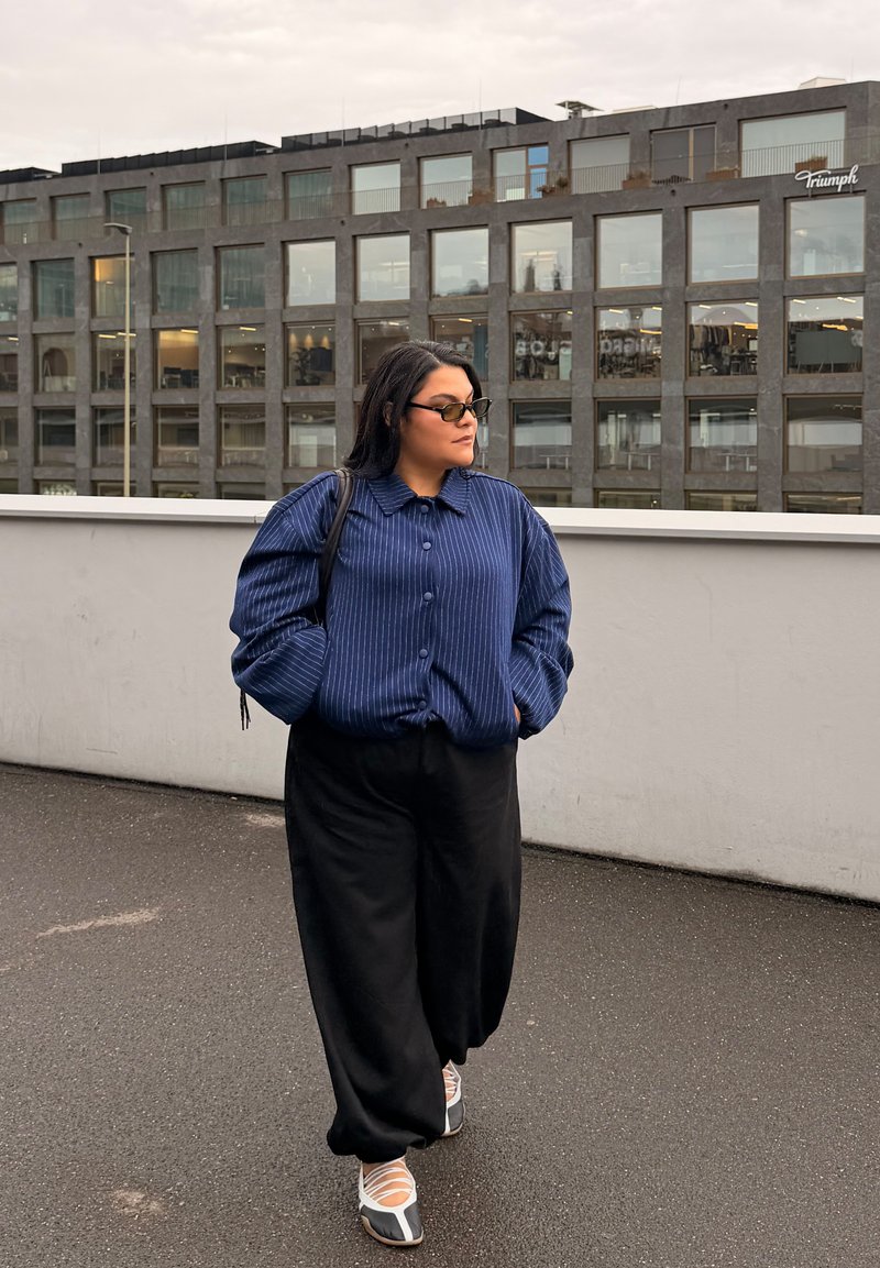 Personne portant des lunettes, une veste rayée bleue et un pantalon noir marchant à l'extérieur à côté d'un bâtiment moderne gris avec de grandes fenêtres en arrière-plan.