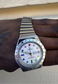 Sidabrinis kvarco Timex laikrodis su apvaliu baltu ciferblatu, daugiaspalviais valandų žymekliais ir nerūdijančio plieno grandininiu dirželiu. Matoma dienos-data funkcija.