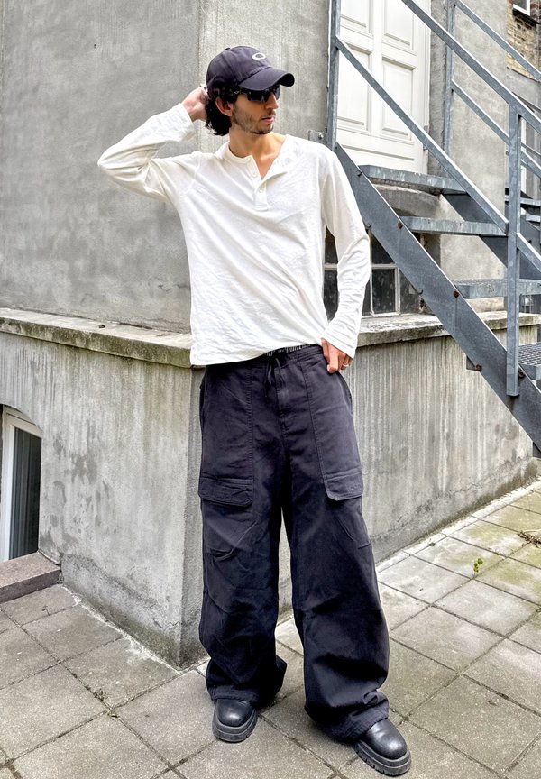 Persona que lleva una camisa blanca de manga larga, pantalones cargo negros, botas, gorra negra y gafas de sol, está de pie al aire libre junto a un edificio de hormigón y una escalera metálica.