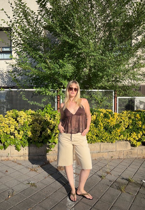 Žena v slnečných okuliaroch, hnedom čipkovanom tope, béžových šortkách a žabkách stojí na chodníku pri zelenom kríku a strome v jasnom slnečnom svetle.