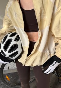 Light beige and cream colour-blocked lightweight jacket with a zip, worn with a black sports bra and brown leggings. Holding a white bicycle helmet.