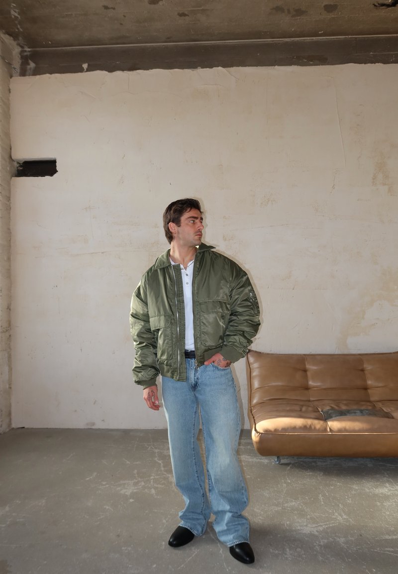 Άνδρας με πράσινο μπουφάν bomber, λευκό πουκάμισο και ανοιχτόχρωμο μπλε τζιν που στέκεται κοντά σε καφέ δερμάτινο καναπέ σε έναν ατελή χώρο.