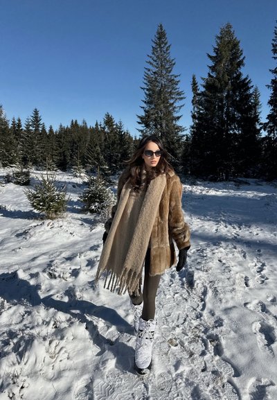 Abrigo de piel sintética marrón, bufanda beige oversized con flecos, leggings oscuros ajustados y botas de nieve blancas con cordones sobre un suelo nevado. Árboles de pino al fondo.